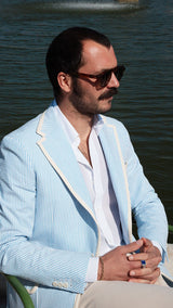 Titouan est assis aux Tuileries porte la veste James en seersucker à rayures bleu ciel et blanc galonnée - Collection Printemps été 2025 - Maison Gabriel Paris