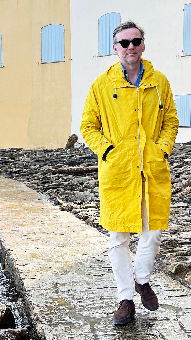Cyril marche dans le quartier de La Ponche à Saint-Tropez et porte le manteau de pluie Jean-Louis en wax jaune - au bord de l'eau - de la Maison Gabriel Paris