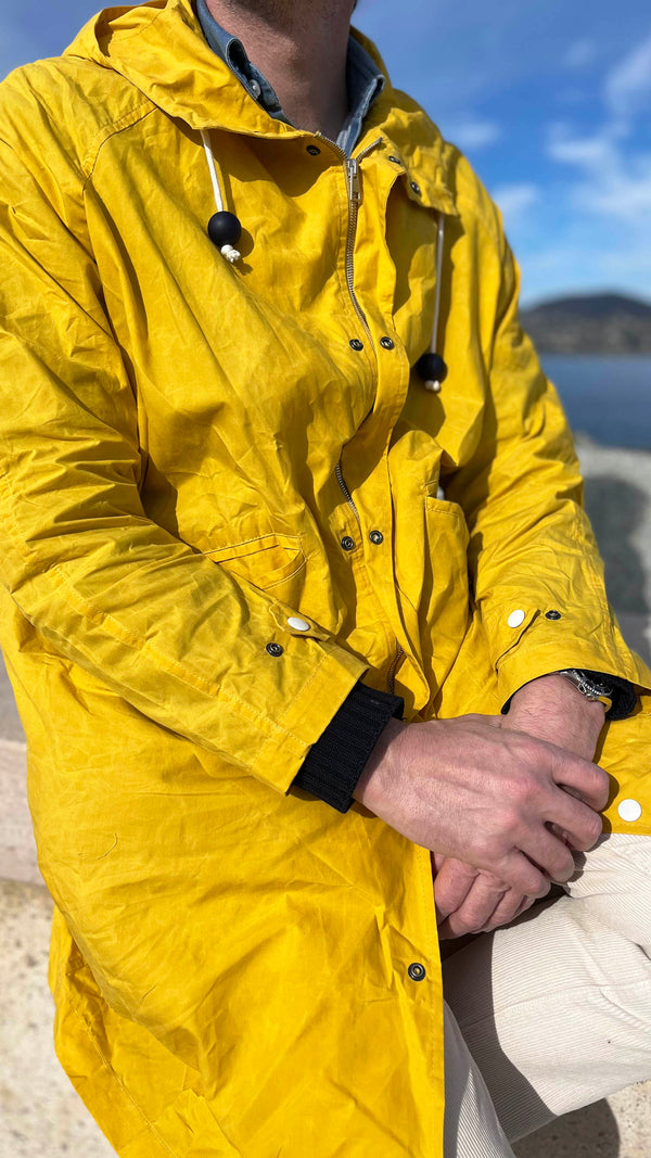 Gros plan du manteau de pluie Jean-Louis en wax jaune porté par Cyril, assis sur un muret à Saint-Tropez Jean-Louis - au bord de l'eau - de la Maison Gabriel Paris