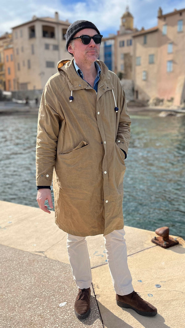 Cyril debout devant la jetée de La Ponche à Saint-Tropez porte le manteau de pluie Jean-Louis en wax kaki - en bord de mer avec le clocher au fond -  de la Maison Gabriel Paris
