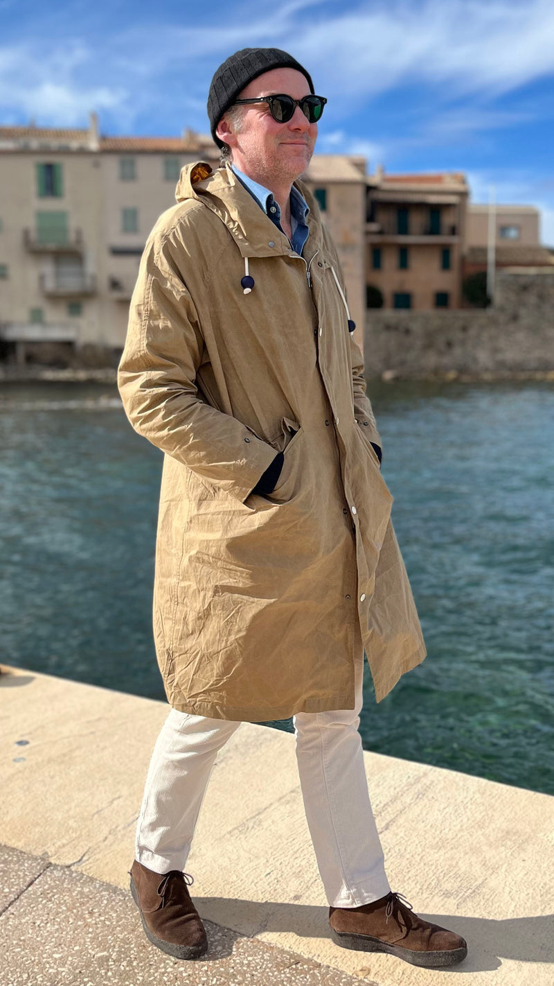 Cyril marche sur la jetée de La Ponche à Saint-Tropez et porte le manteau de pluie Jean-Louis en wax kaki - en bord de mer avec les maisons de pêcheurs au fond -  de la Maison Gabriel Paris