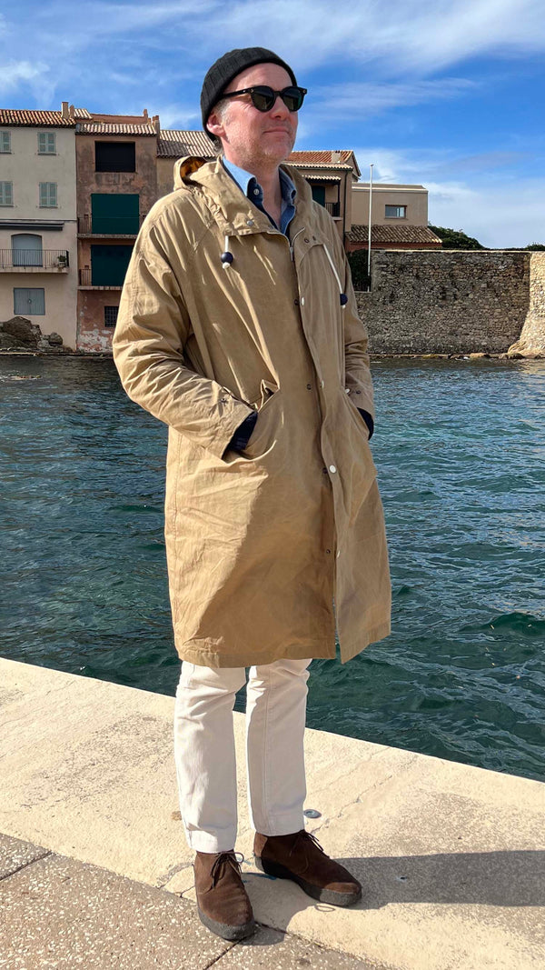 Cyril debout sur la jetée de La Ponche à Saint-Tropez porte le manteau de pluie Jean-Louis en wax kaki - en bord de mer avec les maisons de pêcheurs au fond -  de la Maison Gabriel Paris